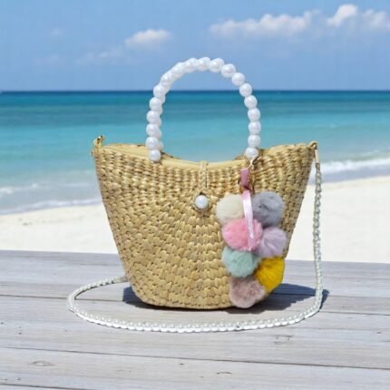 Straw tote bag with pearl handle and pastel pom poms on a beach backdrop